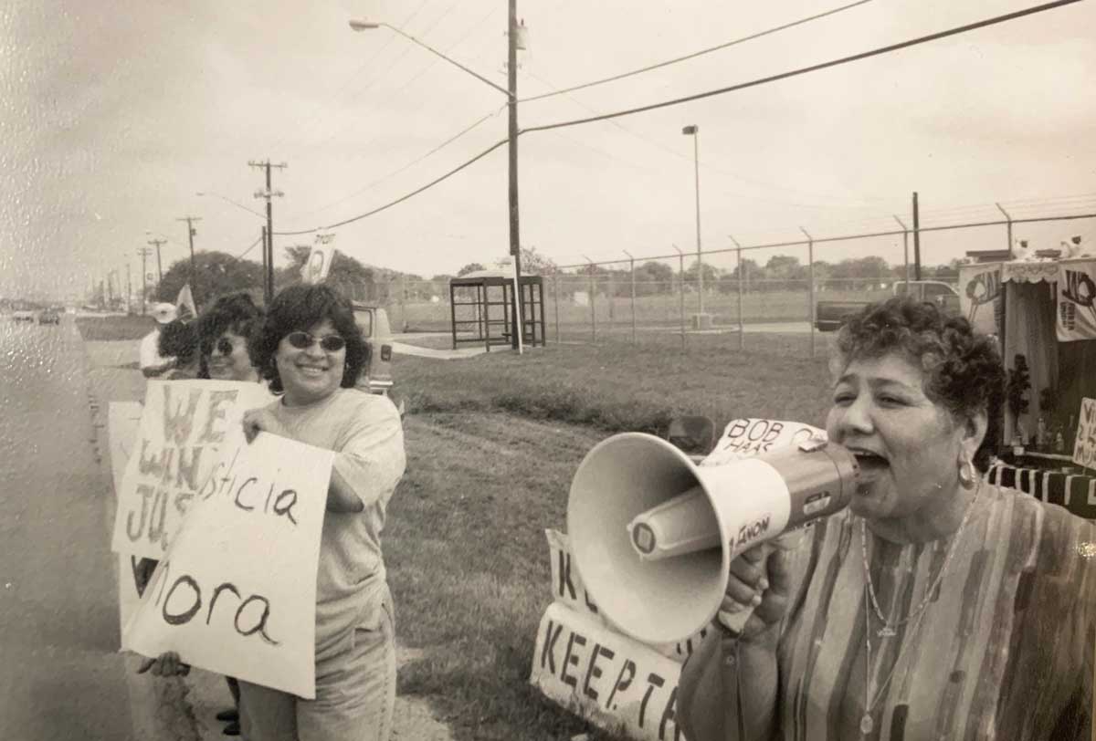A black and white photo shows a female protester speaking into a bullhorn along a road with protest signs and other protesters beside her.