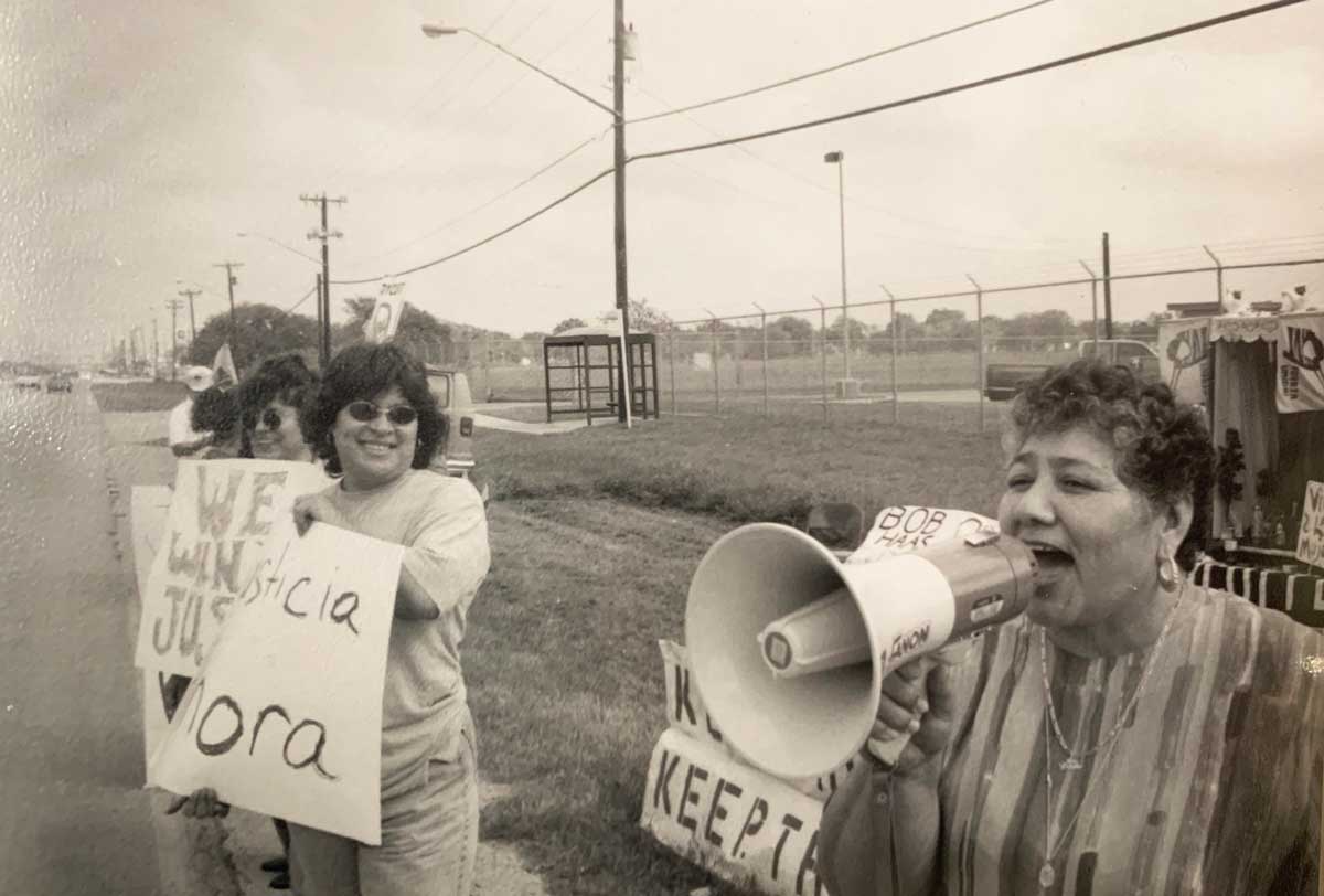 A black and white photo shows a female protester speaking into a bullhorn along a road with protest signs and other protesters beside her. 