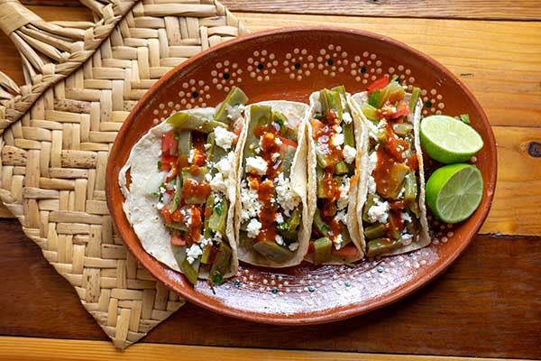 A plate of three tacos with green nopalitos is displayed on a traditional clay Mexican plate.