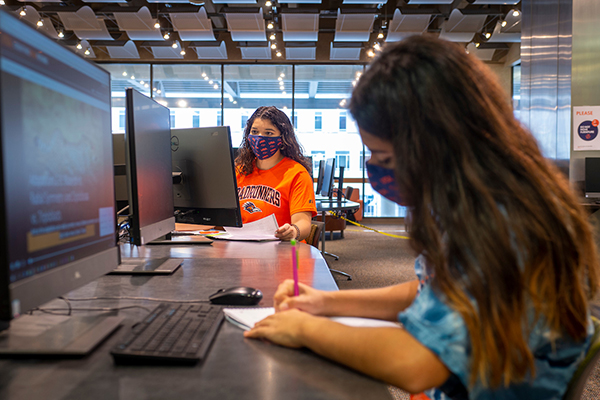 Two students wearing masks study in front of computers at the John Peace Library.