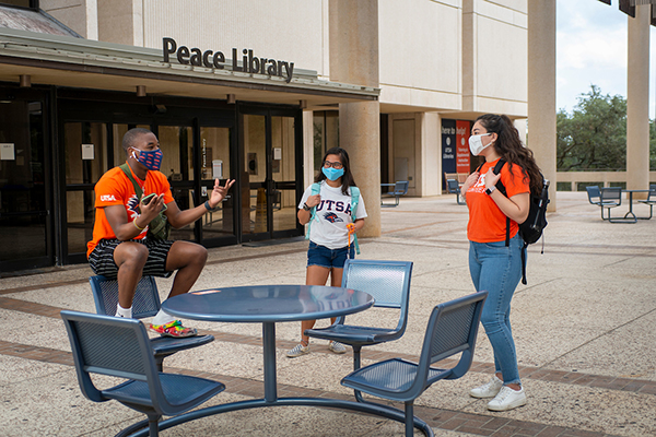 Three UTSA students wearing masks visit with each other outside the second-floor entrance to the John Peace Library.