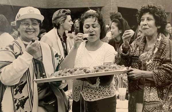 A black-and-white photo shows three women eating food from a platter and smiling.