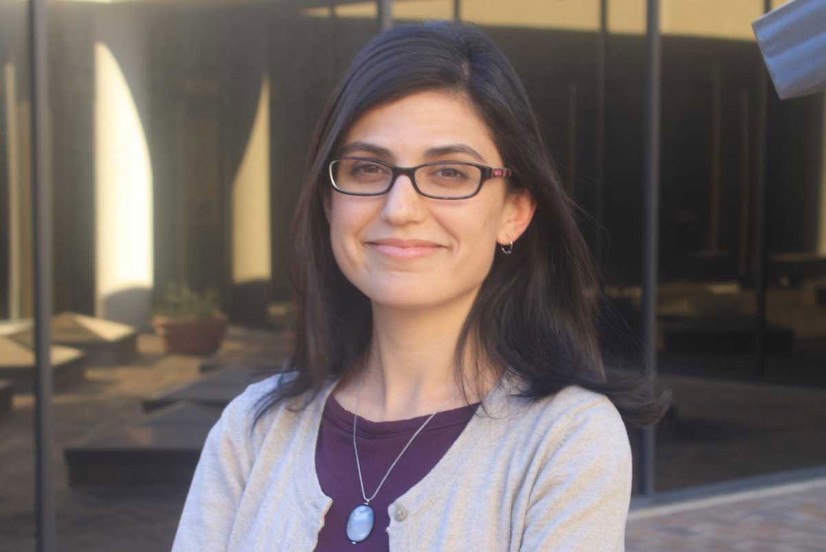 A woman smiles at the camera wearing glasses, a purple blouse and lavender sweater.